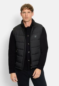 Schwarze gepolsterte Weste mit hohem Kragen, Reißverschluss und Druckknöpfen. Trägt man über einem gerippten schwarzen Pullover mit einem Logo-Patch.