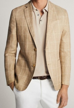 Mand iført en beige ternet blazer over en beige poloshirt, hvide bukser og et brunt flettet bælte, med hænderne i lommerne.