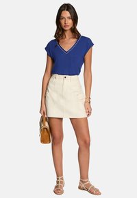 Blauw shirt met korte mouwen en V-hals, gedragen met een crèmekleurige mini-jurk met voorzakken. Gecombineerd met sandalen met bandjes en een tas in de hand.