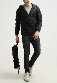 Zwarte pull-over jas met een voorzak, elastische manchetten en een trekkoord-capuchon, gecombineerd met donkere jeans en een zwarte rugzak.