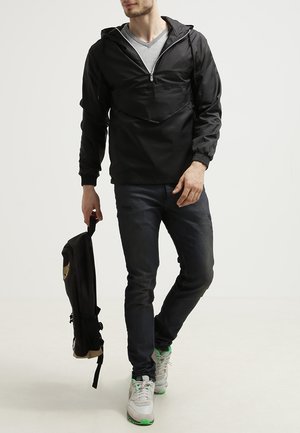 Svart genserjakke med frontlomme, elastiske mansjetter og hette med snor, kombinert med mørke jeans og en svart ryggsekk.