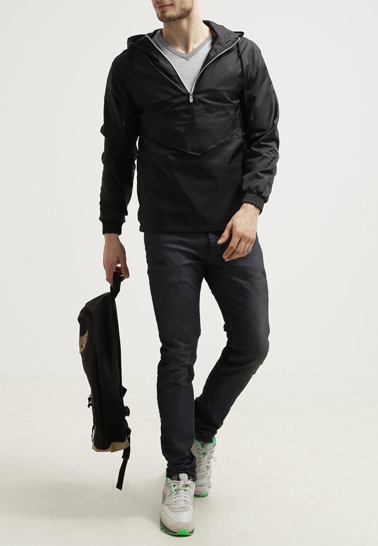 Zwarte pull-over jas met een voorzak, elastische manchetten en een trekkoord-capuchon, gecombineerd met donkere jeans en een zwarte rugzak.