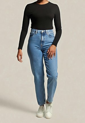 Femme portant un haut noir à manches longues, un jean bleu clair taille haute et des baskets blanches, debout avec une main près de la cuisse.