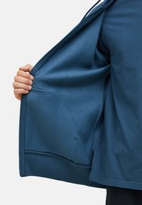 Plava jakna na zip s mekanim, teksturiranim unutrašnjim dijelom, rebrastim rubom i unutarnjim džepom. Čini se da je tkanina topla i izdržljiva.