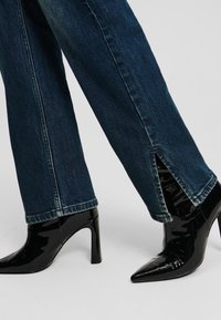 Dark blue jeans with a straight leg and side slit, paired with black patent leather high-heeled boots featuring a pointed toe and sleek design.