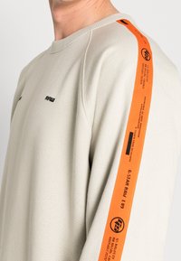 Sudadera beige claro con acabado texturizado, que presenta una franja lateral naranja con texto impreso y cuello redondo.