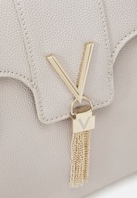 Grå texturerad läderhandväska med en framträdande gyllene "V"-logotyp och gyllene kedjetassel som accent. Har en strukturerad design och sydda kanter.