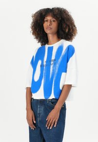 White cropped t-shirt featuring bold blue brushstroke design and black text stating "LOVE IS A FEELING NOT A DECISION." Short sleeves.