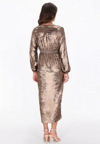 Nő bronzszínű, fémes hatású, hosszú ujjú midi ruhában, húzott derékkal, háttal a kamerának áll, bézs magassarkút visel.