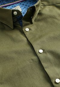 Camisa de botón en verde oliva con botones blancos y un cuello, mostrando un forro interior azul con estampado y una etiqueta de tamaño de la ropa.