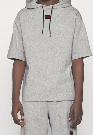 Persoon die een lichtgrijze korte mouwen hoodie draagt met bijpassende korte broek, zwarte trekkoorden en een klein rood "HUGO" logo op de borst.