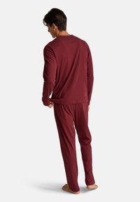 Mężczyzna w bordowej bluzie z długim rękawem i spodniach stoi boso, odwrócony plecami, poprawiając dół swojej bluzki na jednolitym tle.