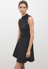 Woman wearing a sleeveless black dress with a high neckline standing against a plain white textured wall.