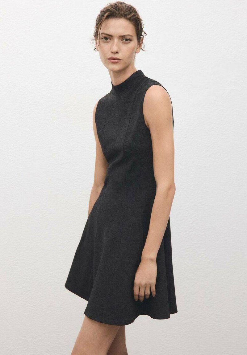 Woman wearing a sleeveless black dress with a high neckline standing against a plain white textured wall.