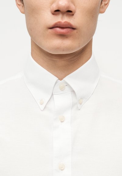 Κοντινό πλάνο ενός νέου άνδρα που φοράει λευκό πουκάμισο με κουμπιά και κουμπωμένο γιακά, μπροστά σε απλό φόντο.