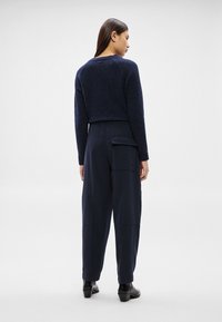 Γυναίκα με την πλάτη γυρισμένη φοράει σκούρο μπλε κοντό πουλόβερ, παντελόνι σε ναυτικό μπλε με ψηλή μέση και μεγάλο πλαϊνό τσέπη, και μαύρα μποτάκια αστραγάλου σε λευκό φόντο.