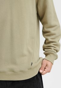 Sudadera beige con puños y dobladillo acanalados. Presenta una textura suave y detalles en las costuras laterales. Etiqueta de la marca pequeña cerca del dobladillo.