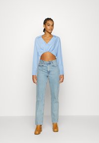 Camisa cropped azul-claro com decote em V e mangas longas, combinada com jeans azul-claro de cintura alta e botas de salto marrons.