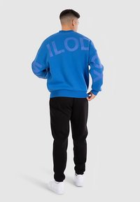 Blå sweatshirt med lila logotypbokstäver, långärmad, ribbade muddar, kombinerad med svarta sweatpants och vita sneakers, fotograferad bakifrån.