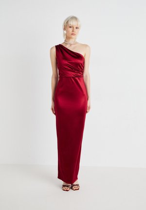 Robe en satin rouge avec une manche, corsage froncé et coupe longue jusqu'au sol. Texture lisse, coupe ajustée, associée à des talons noirs à lanières.