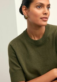 Olivgrüner Kurzarmpullover mit geripptem Ausschnitt. Das Model trägt silberne Ohrringe mit einem großen ovalen Bernsteinstück. Glatte Textur.