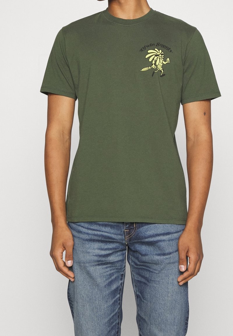 Homme portant un T-shirt vert olive avec un petit personnage de dessin animé jaune et le texte "TAVIN SUPPLY" sur le côté gauche de la poitrine, associé à un jean bleu.