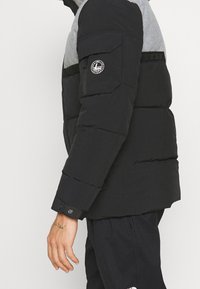 Chaqueta de plumas negra con sección superior gris, que presenta un parche de logo en la manga, bolsillo con cremallera y puños ajustables. Textura suave.