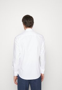 Biała koszula z długim rękawem, z zaokrąglonym dołem, mankietami na guziki i gładką fakturą, noszona przez postać widzianą od tyłu na jednolitym tle.