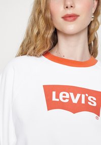 Fehér pulóver narancssárga bordázott gallérral és nagy piros Levi's logóval a közepén. Puha anyag és laza szabású dizájn.