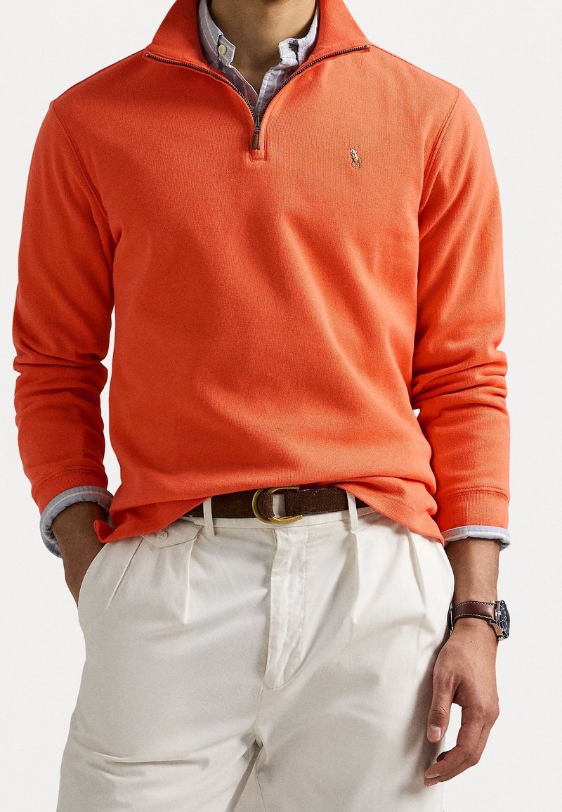 Homme portant un pull orange à fermeture éclair jusqu'au quart, par-dessus une chemise bleu clair rentrée dans un pantalon blanc plissé avec une ceinture marron et une montre-bracelet.