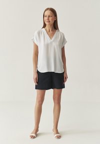 Blusa blanca de manga corta con escote en V, combinada con pantalones cortos azul marino. Textura suave, corte relajado y detalle de dobladillo redondeado.
