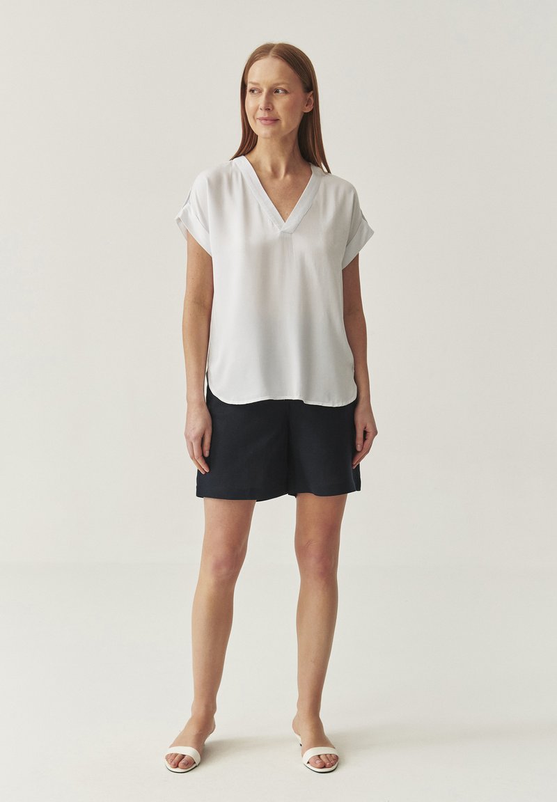 Blusa blanca de manga corta con escote en V, combinada con pantalones cortos azul marino. Textura suave, corte relajado y detalle de dobladillo redondeado.