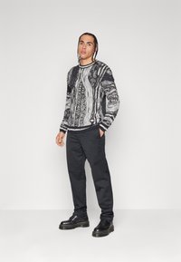 Schwarzer und weißer gemusterter Strickpullover mit langen Ärmeln, kombiniert mit schwarzen Hosen und schwarzen Schuhen mit dicken Sohlen.