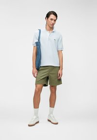 Chemise polo à manches courtes bleu clair, shorts vert olive, baskets blanches avec semelles brunes et un sac en toile en denim.