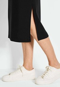 Schwarzes Midikleid mit seitlichen Schlitzen, festem Saum und weichem Stoff, kombiniert mit weißen Sneakers mit abgerundeter Spitze und flacher Sohle.