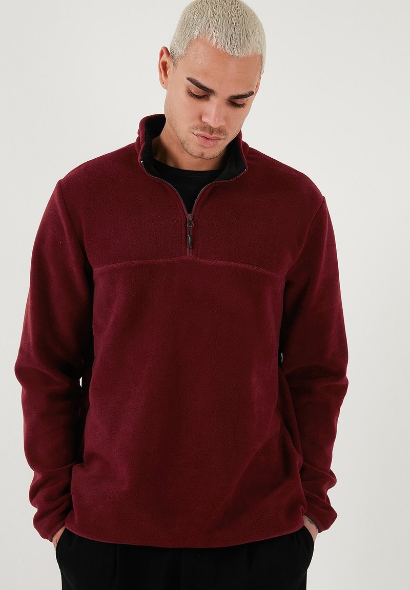 Pullover in pile color bordeaux con colletto alto, mezza zip e dettagli neri. Texture morbida, maniche lunghe e cucitura orizzontale.
