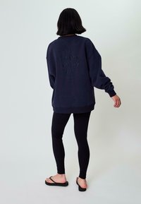 Felpa blu navy oversize con ricamo testurizzato che recita "STO CREANDO LA MIA MAGIA" sul retro, abbinata a leggings neri e infradito.