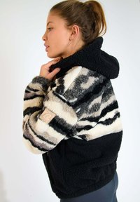 Giacca in pile nera, beige e grigia con un motivo a strisce testurizzato. Presenta un cappuccio e una zip laterale, e una toppa con logo sulla manica.