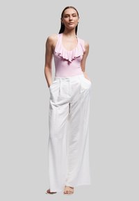 Roze halter-nek badpak met gerafelde details, gecombineerd met witte, hoge taille, geplooide broek. Model staat met handen in de zakken.