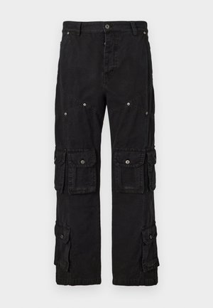 Homme portant des lunettes de soleil noires, un t-shirt gris, une veste zippée à carreaux foncés, un pantalon cargo noir et des baskets noir et blanc, debout avec une main dans la poche.