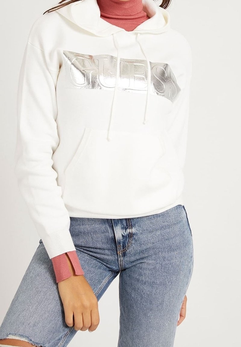 Sweat à capuche blanc avec logo Guess argenté, porté sur un col roulé rose, associé à un jean bleu clair déchiré sur une personne debout.