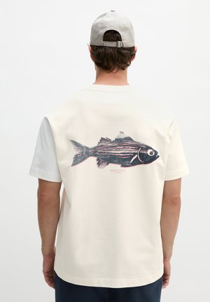 Homme portant un t-shirt blanc avec un grand motif de poisson dans le dos et une casquette gris clair, de dos sur un fond uni.