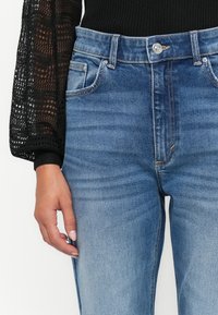 Svetlo modre jeans hlače z visokim pasom, rahlo zbledelimi in petimi žepi. Imajo kovinsko gumb in kontrastno šivanje.