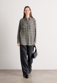 Camicia button-up a quadri grigia con due tasche sul petto, abbinata a pantaloni larghi scuri e una borsa nera. Il modello è in piedi su uno sfondo neutro.