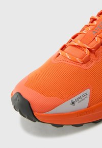 Oranžová běžecká obuv s texturovaným syntetickým svrškem, polstrovaným límcem a štítkem GORE-TEX. Disponuje černou gumovou podrážkou a brandingem LIGHTSTRIKE.