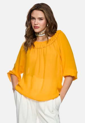 Woman with wavy brown hair wearing a yellow ruffled blouse, white pants, and a silk neck scarf, standing with hands in pockets.