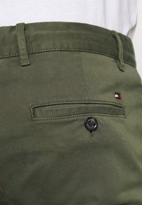 Groene katoenen broek met een achterzak voorzien van een zwarte knop en subtiele stiksel. Een klein rood, wit en blauw logo is zichtbaar nabij de zak.