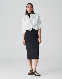 Weißes Hemd mit gebundenen Knöpfen über einem schwarzen strukturierten Midirock, kombiniert mit schwarzen Sandalen. Das Outfit zeichnet sich durch ein entspanntes, geschichtetes Design aus.