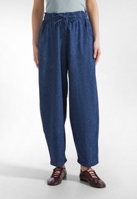 Osoba ubrana w luźne, ciemnoniebieskie spodnie z elastycznym pasem i sznurkiem, w parze z bordowymi, wiązanymi płaskimi butami.