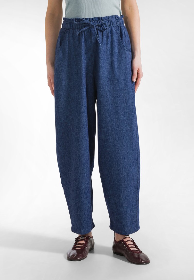 Osoba ubrana w luźne, ciemnoniebieskie spodnie z elastycznym pasem i sznurkiem, w parze z bordowymi, wiązanymi płaskimi butami.